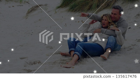 Pointing mature couple wearing jeans and sweaters cuddling on sandy dunes at beach, copy space 135617476