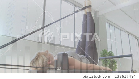 Lying woman wearing leggings extending legs on Pilates reformer in fitness studio, with foot straps Lying woman wearing leggings extending legs on Pilates reformer in fitness studio, with foot straps 135617484