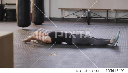 Lying woman wearing workout gear stretching on rubber mats at gym with punching bags and barbell 135617491