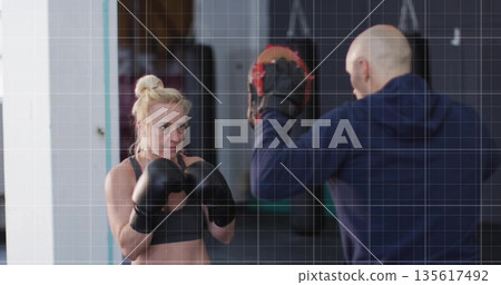 Trainee wearing sports bra punching focus mitts with boxing gloves in boxing gym, copy space Trainee wearing sports bra punching focus mitts with boxing gloves in boxing gym, copy space 135617492
