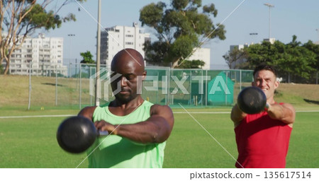 Swinging kettlebells man in green tank on sports field, with chain-link fence and red-shirt partner Swinging kettlebells man in green tank on sports field, with chain-link fence and red-shirt partner 135617514