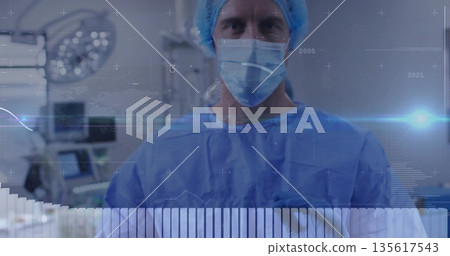 Standing male surgeon wearing blue gown and mask in operating room, with monitors and overlays Standing male surgeon wearing blue gown and mask in operating room, with monitors and overlays 135617543