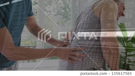 Younger caregiver massaging senior client back in therapy room with plant, window and code overlay Younger caregiver massaging senior client back in therapy room with plant, window and code overlay 135617565