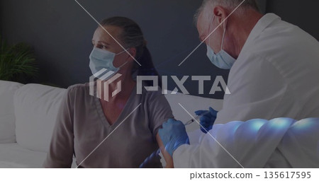 Doctor injecting vaccine into patient arm on couch at home, with syringe and gloves 135617595