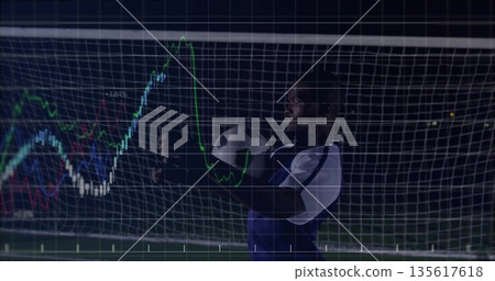 Goalkeeper gripping soccer ball before goal net on floodlit field, with digital line graphs overlay 135617618
