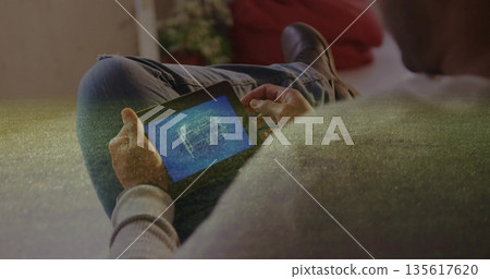 Seated man wearing grey sweater reviewing blueprint on tablet in living room, with red cushion 135617620