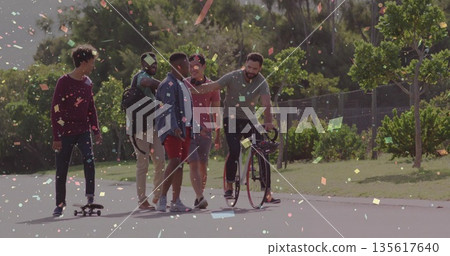 Walking five friends chatting along tree-lined park path amid confetti with bicycle and skateboard Walking five friends chatting along tree-lined park path amid confetti with bicycle and skateboard 135617640