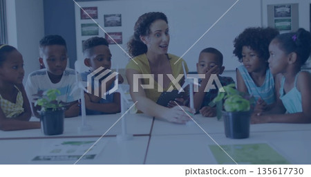 Gesturing teacher in yellow blouse explaining wind turbine models at classroom table, with tablet 135617730