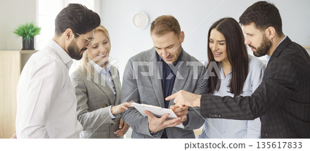 Smiling business team standing in office, exchanging ideas and discussing document during meeting. Smiling business team standing in office, exchanging ideas and discussing document during meeting. 135617833