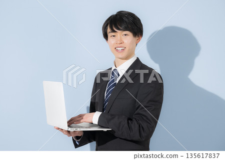 A young male university student in a suit holding a laptop on a light blue background. Business image of new graduates and job hunting. Recruitment image. A young male university student in a suit holding a laptop on a light blue background. Business image of new graduates and job hunting. Recruitment image. 135617837