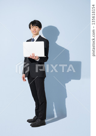 A young male university student in a suit holding a laptop on a light blue background. Business image of new graduates and job hunting. Full body image of job hunting recruitment. A young male university student in a suit holding a laptop on a light blue background. Business image of new graduates and job hunting. Full body image of job hunting recruitment. 135618154