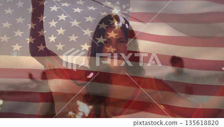 Holding sparkler above head woman wearing red top on beach at dusk, with American flag overlay Holding sparkler above head woman wearing red top on beach at dusk, with American flag overlay 135618820
