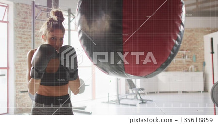 Woman boxer wearing sports bra throwing punches at punching bag in gym studio, with boxing gloves 135618859