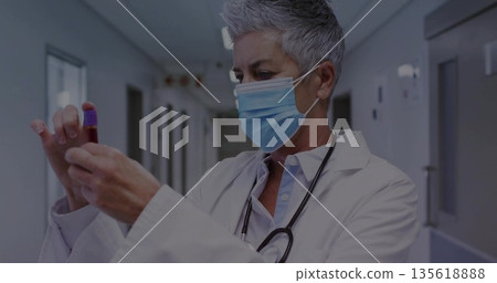 Senior doctor wearing mask inspecting blood sample vial in hospital corridor, with stethoscope 135618888
