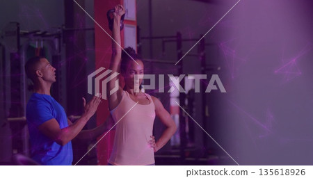 Woman lifting kettlebell overhead in gym area with trainer and squat racks barbells weight plates 135618926