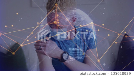 Man wearing blue t-shirt and medical face mask holding left shoulder on couch, with network overlay 135618929