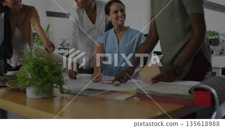 Reviewing women designers examining blueprints at table in workspace, with swatches and plant 135618988