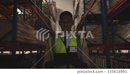 Warehouse inspector scanning pallets on metal racks in narrow aisle, with tablet and safety vest Warehouse inspector scanning pallets on metal racks in narrow aisle, with tablet and safety vest 135618991