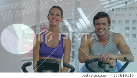 Pedaling smiling woman and man in sportswear on stationary bikes in gym, with translucent graphs 135619116