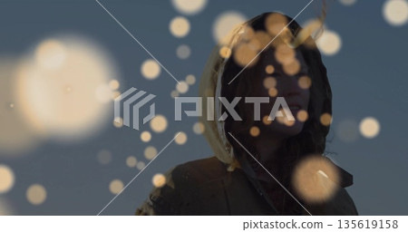 Standing adult female wearing hooded parka outdoors against dusk sky, with warm-toned bokeh overlay Standing adult female wearing hooded parka outdoors against dusk sky, with warm-toned bokeh overlay 135619158