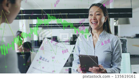 Smiling Korean woman holding tablet and discussing financial charts in office, with monitors 135619161