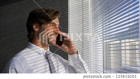 Talking businessman wearing shirt tie holding smartphone beside office window blinds, copy space Talking businessman wearing shirt tie holding smartphone beside office window blinds, copy space 135619201