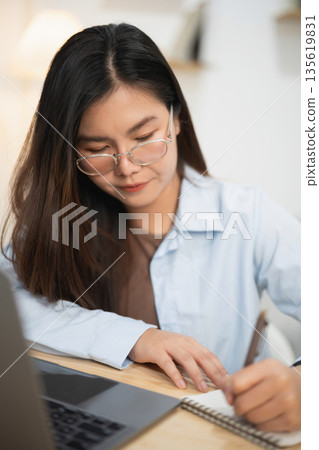 Young woman focused on writing notes while using laptop in bright modern workspace with soft lighting, showcasing productivity and concentration Young woman focused on writing notes while using laptop in bright modern workspace with soft lighting, showcasing productivity and concentration 135619831