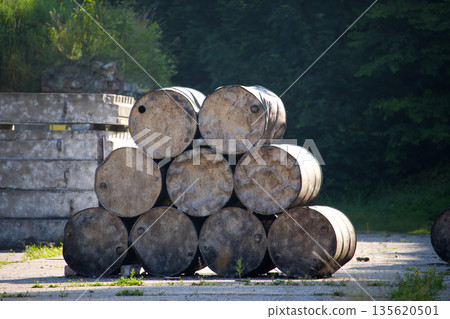 A pyramid of old, weathered metal barrels rests on a concrete surface under sunlight. A pyramid of old, weathered metal barrels rests on a concrete surface under sunlight. 135620501