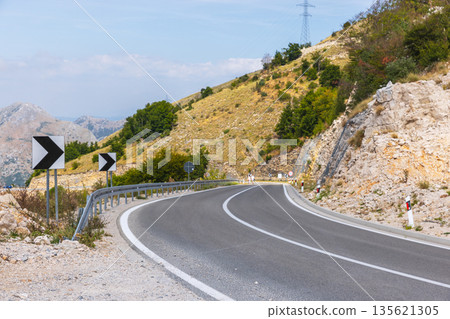 A winding mountain road bends through rocky hills with guardrails and chevron signs A winding mountain road bends through rocky hills with guardrails and chevron signs 135621305