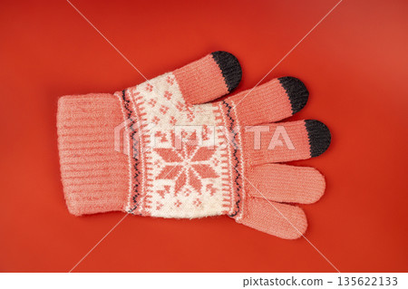 Pink knitted winter glove with white snowflake pattern on a red background. Warm seasonal accessory. Pink knitted winter glove with white snowflake pattern on a red background. Warm seasonal accessory. 135622133