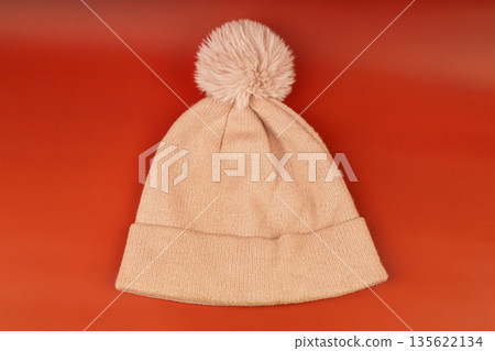 Pink knitted winter hat with a pompom on a red background. Warm wool beanie for cold weather. 135622134