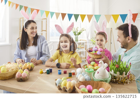Happy family wearing bunny ears, decorating colorful eggs together at home in bright festive room. 135622715