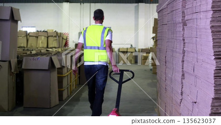 Warehouse worker wearing yellow vest pushing red pallet jack past cardboard boxes and pallets Warehouse worker wearing yellow vest pushing red pallet jack past cardboard boxes and pallets 135623037