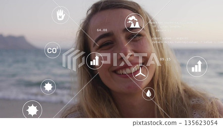 Smiling blonde woman standing on beach with digital overlays showing CO2 icon, other icons and code 135623054