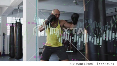 Punching man wearing yellow top and gloves delivering punch at gym, with heavy bag, chart overlays 135623074