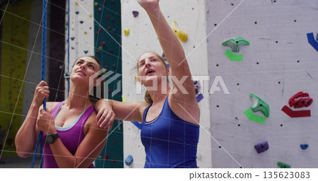 Pointing instructor guiding belaying climber at climbing gym wall, with rope harness and handholds 135623083