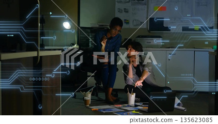 Colleagues discussing floor-laid design plans in dim workspace, with laptop overlay Colleagues discussing floor-laid design plans in dim workspace, with laptop overlay 135623085