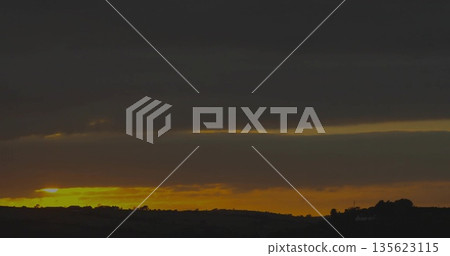 Glowing yellow orange horizon strip spanning rolling hills in countryside at dusk, moody cloudy sky 135623115