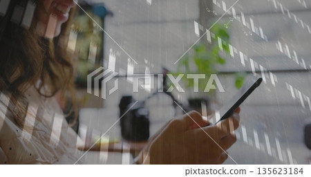 Woman wearing polka-dot blouse tapping smartphone in home office, with camera, plant, data charts 135623184