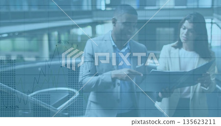 Standing professionals wearing suits examining binder by glass railing in atrium, with charts 135623211