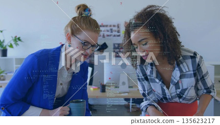 Leaning over desk two female coworkers in blazers examining code in office, with ceramic coffee mug 135623217