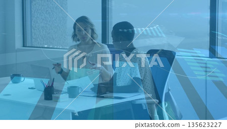Two businesswomen discussing documents while sitting at desk in office with laptop and coffee mugs Two businesswomen discussing documents while sitting at desk in office with laptop and coffee mugs 135623227