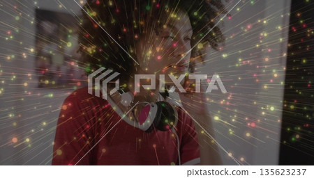 Standing woman wearing red T-shirt, over-ear headphones by wall, with bursting colorful particles 135623237