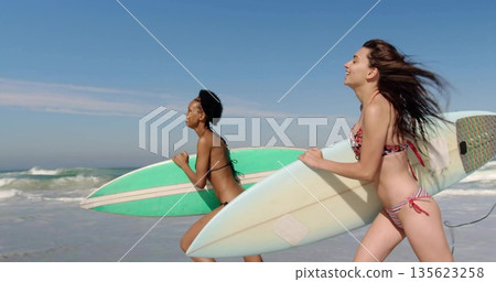 Sprinting pair in bikinis with surfboards across beach with green-striped and pale-patterned boards Sprinting pair in bikinis with surfboards across beach with green-striped and pale-patterned boards 135623258