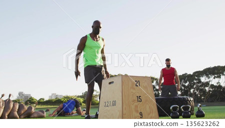 Working out man in green tank top beside plyometric box on park grass, with fitness equipment 135623262