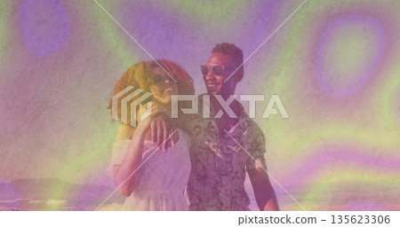 Walking couple strolling sunlit ocean shoreline, with sunglasses summer dress and patterned shirt 135623306