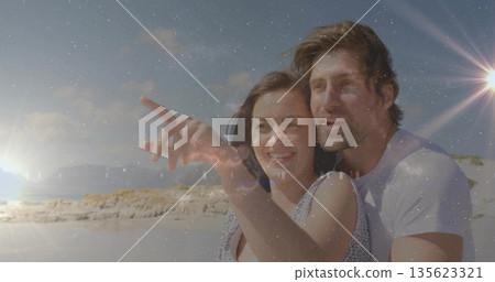 Pointing male couple wearing casual beachwear embracing on sandy shoreline, with warm sunset glow Pointing male couple wearing casual beachwear embracing on sandy shoreline, with warm sunset glow 135623321