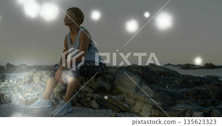 Sitting woman in athletic clothing gazing upward on rocky beach, with glowing orbs, copy space Sitting woman in athletic clothing gazing upward on rocky beach, with glowing orbs, copy space 135623323
