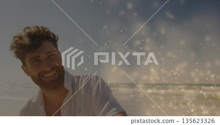 Smiling bearded man wearing white shirt standing on shoreline, sparkling light overlay, copy space 135623326