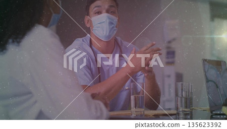 Speaking doctor in scrubs and face mask at clinic desk, with stethoscope laptop papers water Speaking doctor in scrubs and face mask at clinic desk, with stethoscope laptop papers water 135623392
